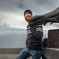 Ein lächelnder Mann mit rotem Vollbart lehnt entspannt an einem Geländer an einem windigen Strand. Er trägt eine dunkle Wollmütze, einen dicken Pullover mit skandinavischem Muster und eine Jeans. Eine grüne Jacke wird vom Wind hochgeweht und flattert dramatisch in der Luft.