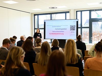 Heller Seminarraum mit großen Fenstern und Blick nach draußen. Eine Referentin steht vorne neben einem großen Bildschirm und präsentiert vor einem sitzenden Publikum. Auf der Folie sind Stichworte wie „Wetterdaten“, „Marketingdaten“, „Besucherzahlen“ und „Kundenbestimmung“ sowie ein QR-Code zu sehen.