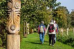 Eine Gruppe Menschen wandert auf einem grünen Wiesenpfad durch eine naturnahe, waldreiche Landschaft. Links im Vordergrund steht eine große, kunstvoll geschnitzte Holzskulptur in Form einer stilisierten Eule. Weitere geschnitzte Figuren und Pfosten mit gelben Markierungen säumen den Weg. 