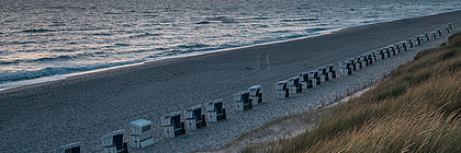Blick von der grün bewachsenen Düne aus über den Strand von Rantum, auf dem eine lange Reihe von weißen Strandkörben steht. Im Mittelgrund die Wellen der Nordsee, im Hintergrund ein leicht bewölkter Abendhimmel. Am Horizont rot-gelbe Himmelsfärbung. - zum Artikel 'Die aktuelle Buchungslage im Überblick'