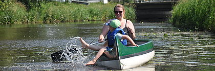 Eine Frau in einem grünen Kanu paddelt durch die Wilsterau im Holsteiner Auenland in der Nähe von Itzehoe. Vorn im Kanu sitzt ein Kind und planscht mit den Füßen im Wasser.  - zum Artikel 'Wassersportparadiese in Schleswig-Holstein: Unterwegs mit Paddel & Co. '