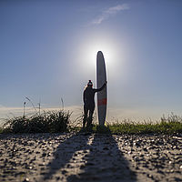 Surfer/in steht mit Surfbrett am Strand mit Blick Richtung Sonne