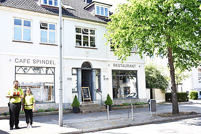 Eine helle Straßenansicht zeigt die Fassade eines Gebäudes mit der Aufschrift „Café Spindel“ auf der linken Seite und „Restaurant“ auf der rechten Seite. Dazwischen befindet sich ein Eingang mit dunkler Tür, beidseitigen Fenstern und einem kleinen Treppenaufgang. Ein Holzschild mit Tagesangebot steht vor dem Eingang. Links im Bild stehen zwei Personen.