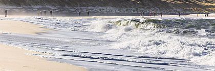 Urlauber spazieren am Strand des Sylter Ellenbogens bei tosender Nordseebrandung und strahlendem Herbstsonnenschein. Im Hintergrund ragt die Spitze des Leuchtfeuers List-West hervor - zum Artikel 'Buchungslage im Schleswig-Holstein-Tourismus für Herbst mit Ausblick auf Winter 2023'