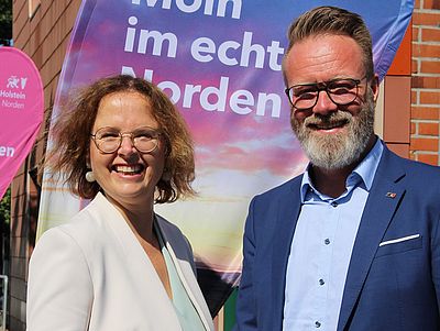 Dr. Bettina Bunge und Claus Ruhe Madsen stehen nebeneinander im Freien vor einem Banner mit der Aufschrift „… im echten Norden“. Beide lächeln in die Kamera.