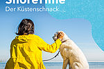 Eine Frau sitzt mit einem gelben Regenmantel bekleidet auf dem Sandstrand am Meer. An der Leine führt sie einen weißen Hund. Beide wirken entspannt und gucken aufs blaue Meer hinaus.