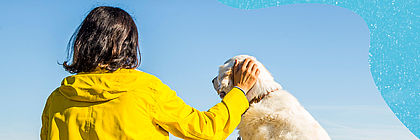 Eine Frau sitzt mit einem gelben Regenmantel bekleidet auf dem Sandstrand am Meer. An der Leine führt sie einen weißen Hund. Beide wirken entspannt und gucken aufs blaue Meer hinaus. - zum Artikel 'Gut vorbereitet und bester Laune: Urlaub mit Hund im echten Norden'
