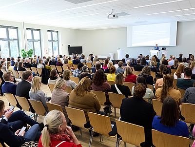 Großer, heller Veranstaltungssaal mit vielen Reihen von Holzstühlen, die dicht besetzt sind. Zahlreiche Menschen sitzen und blicken nach vorne zu einer Bühne. Dort steht Staatssekretärin Julia Carstens am Rednerpult und hält einen Vortrag, während hinter ihr eine Präsentation auf eine Leinwand projiziert wird.