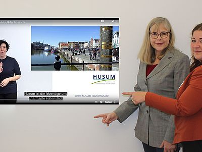 Zwei Frauen stehen vor einer weißen Wand und zeigen lächelnd auf ein eingeblendetes YouTube-Video. Auf dem Bildschirm ist ein Video mit dem Titel „Husum in 90 Sekunden in Gebärdensprache“ zu sehen. Links im Video steht eine Frau mit dunklem Haar, die Gebärdensprache nutzt.