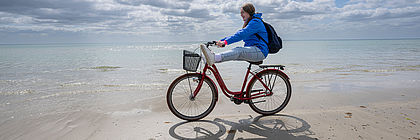 Eine junge Frau fährt auf einem roten Fahrrad durch den Sand an der Ostsee. Sie lacht und legt die Beine beim Fahren auf den Lenker. Im Hintergrund ist die Ostsee zu sehen sowie ein paar Wolken am hellblauen Himmel. - zum Artikel 'Weltfahrradtag: Abenteuer für Fahrradfans in Schleswig-Holstein '