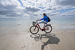 Eine junge Frau fährt auf einem roten Fahrrad durch den Sand an der Ostsee. Sie lacht und legt die Beine beim Fahren auf den Lenker. Im Hintergrund ist die Ostsee zu sehen sowie ein paar Wolken am hellblauen Himmel.
