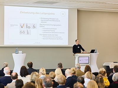 Bosse Willenberg steht an einem Rednerpult in einem großen, hellen Veranstaltungssaal und hält einen Vortrag vor einem sitzenden Publikum. Neben ihm stehen ein Laptop sowie Wasserflaschen und Gläser auf einem Stehtisch. Hinter ihm ist eine große Präsentationsfolie mit dem Titel „Zielsetzung des Leitprojekts“ zu sehen, die Stichpunkte und Symbole zum Thema Barrierefreiheit zeigt.