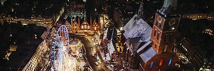 Luftaufnahme vom Maritimen Weihnachtsmarkt auf dem Koberg in Lübeck. Der Weihnachtsmarkt ist hell erleuchtet bei dunklem Himmel. Neben einem Riesenrad ist links die Jakobikirche zu sehen. Es liegt wenig Schnee auf den Dächern. - zum Artikel 'Zauberhafte Weihnachtsmärkte in Schleswig-Holstein '