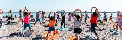 Gruppe macht Yoga am Strand in Schleswig-Holstein bei Sonnenschein – gemeinsames Training mit Blick aufs Meer im Urlaub. - zum Artikel 'Buchungslage für die Sommerferien 2025 in Schleswig-Holstein'