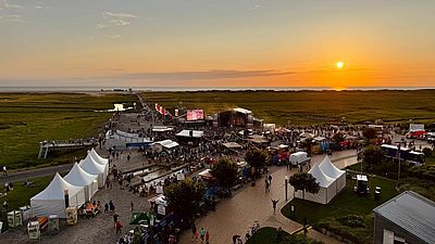 Open-Air-Veranstaltung mit Bühne und Publikum bei Sonnenuntergang an der Nordseeküste, umgeben von Wiesen, Zelten und Verkaufsständen.