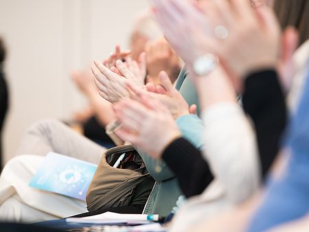 Mehrere Personen sitzen in Reihen und applaudieren. Der Fokus liegt auf den klatschenden Händen im Vordergrund, während die Gesichter unscharf und größtenteils nicht sichtbar sind.