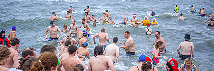 Neujahrsanbaden in Büsum. Viele Menschen in bunter Badekleidung und einigen nostalgischen Badekappen laufen ins Meer. Ein Mann in einem roten Taucheranzug sichert das Anbaden an der Küste vor Büsum und steht zwischen den mutigen Anbadern.  - zum Artikel 'Festtagsflucht: Kurz mal raus an den Feiertagen'