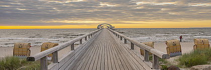 Die Seebrücke von Kellenhusen steht im Zentrum des Bildes. Die Blickrichtung entlang der Brücke führt über den Strand mit Strandkörben und das Meer bis zum bewölkten Horizont.  - zum Artikel 'Verhalten positive Sommerbilanz für Schleswig-Holstein'
