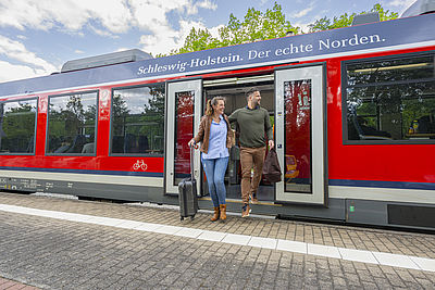 Zwei Personen steigen mit Gepäck aus einem roten Regionalzug an einem Bahnhof aus. Die Frau zieht einen Rollkoffer hinter sich her, der Mann trägt eine Reisetasche. Über den Türen des Zuges steht der Schriftzug „Schleswig-Holstein. Der echte Norden.“.
