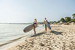 Eine Frau und ein Mann in Badebekleidung laufen mit SUP-Boards und Paddel am Strand entlang.