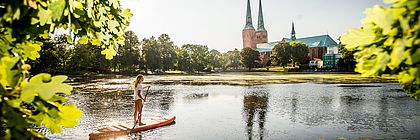 Eine Person fährt auf einem SUP auf dem Mühlenteich. Im Hintergrund ist der Dom zu Lübeck zu sehen.  - zum Artikel 'Willkommen zum DZT Incoming & Brand Summit in Lübeck'