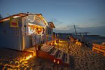 Eine beleuchtete Beachbar am Strand. Eine Frau wird gerade an der Bar bedient, während Personen auf Palletten-Stühlen davor sitzen.
