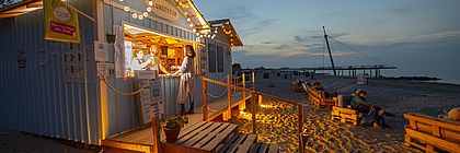Eine beleuchtete Beachbar am Strand. Eine Frau wird gerade an der Bar bedient, während Personen auf Palletten-Stühlen davor sitzen. - zum Artikel 'Das ist neu an der Ostsee: Hoteleröffnungen, neue Restaurants und frische Freizeitangebote'